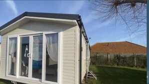 A small, single-story mobile home with a pitched roof, large windows, and a sliding door. - Impeccable 3-bed Caravan in Clacton-on-sea (Clacton-on-Sea)