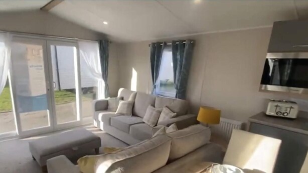 A living room with a sofa, a coffee table, and a TV. There is a sliding glass door leading to an outdoor area. - Impeccable 3-bed Caravan in Clacton-on-sea (Clacton-on-Sea)
