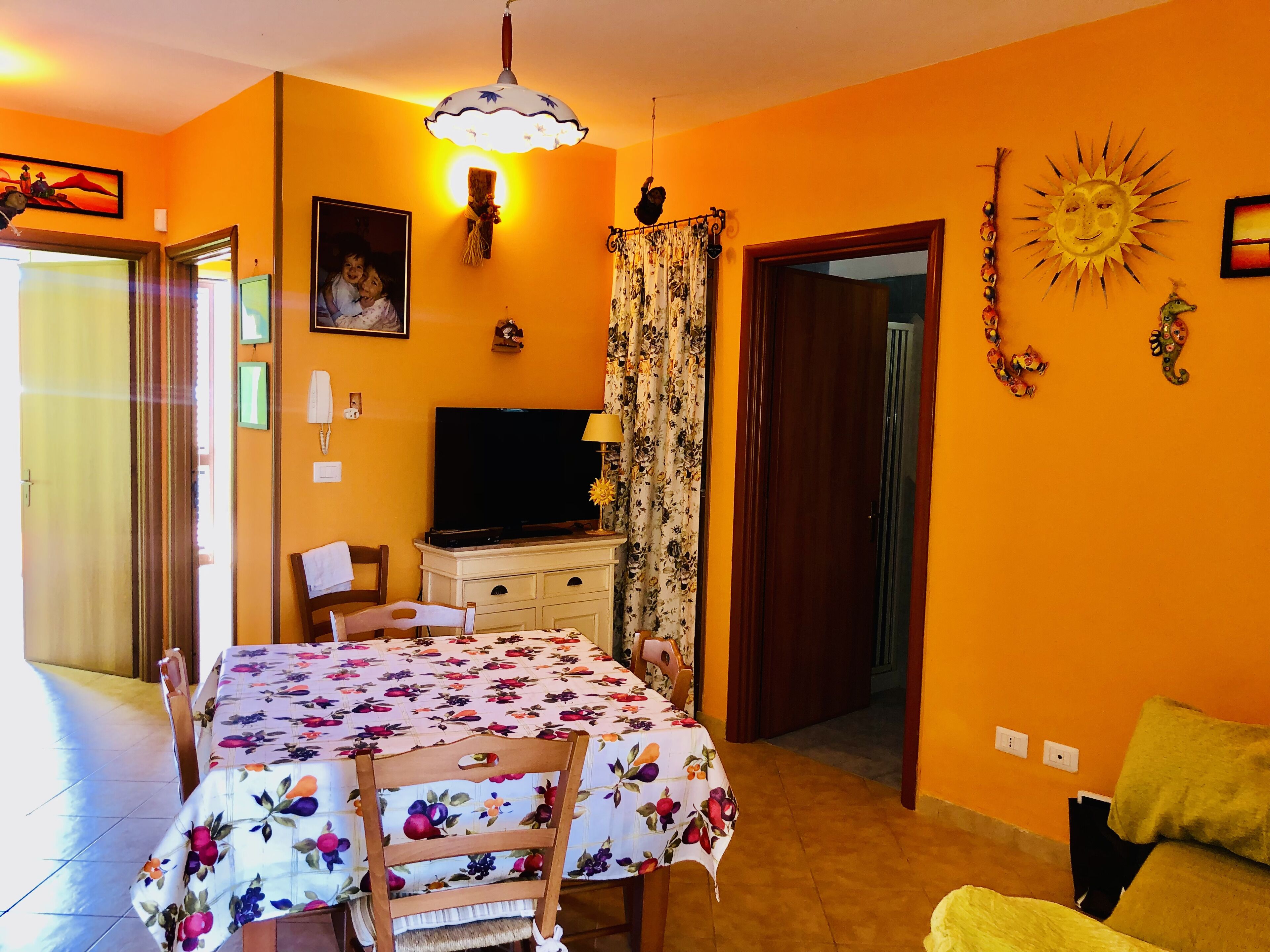 A dining area with a table covered in a floral tablecloth, chairs, a television, and a decorative sun on the wall.