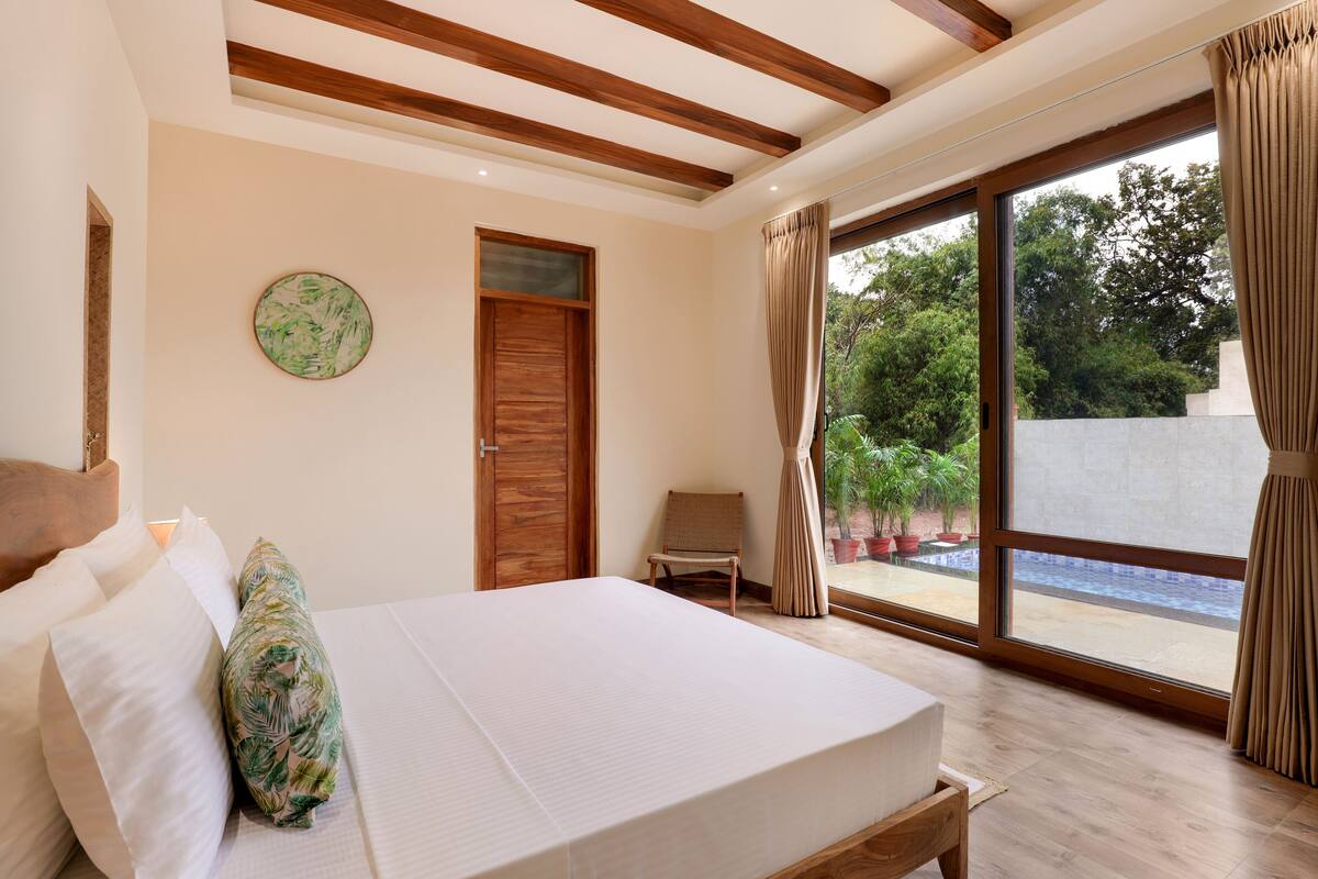 A bedroom with a bed, a wooden door, a chair, and a view of a pool through large windows.