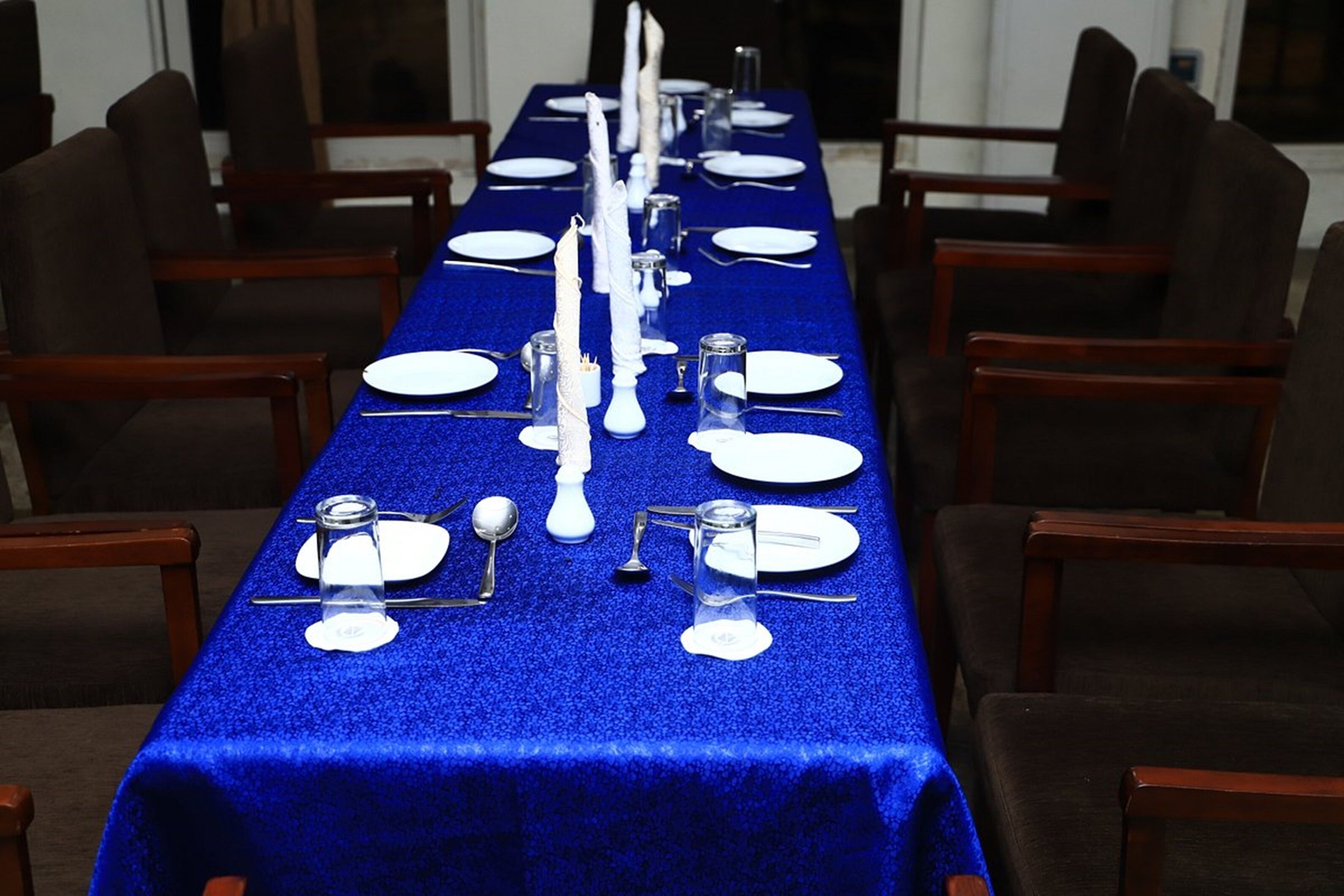 Una larga mesa de comedor dispuesta con platos, cubiertos y vasos, rodeada de sillas.