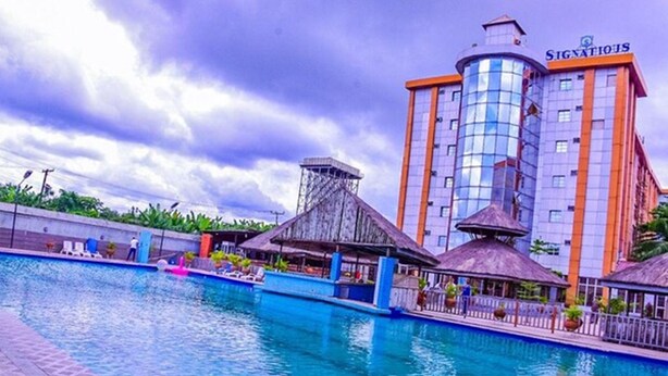 A hotel with a swimming pool, a thatched-roof structure, and a tall building with the sign "SIGNATIOUS". - Signatious Hotel and Suites (Warri)