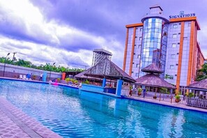 Un hotel con piscina, una estructura de techo de paja y un edificio alto con el letrero "SIGNATIOUS".