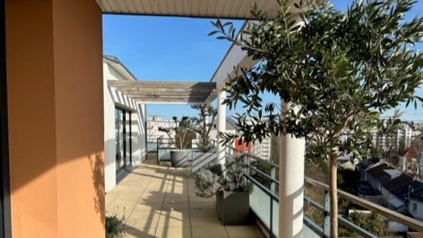 Ein überdachter Balkon mit einem Blumentopf, einer Sitzecke und Blick auf die Stadtlandschaft.