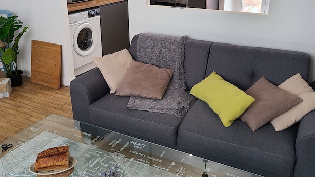 Un salon moderne avec un canapé gris, une table basse en verre et un coin cuisine à l’arrière-plan.