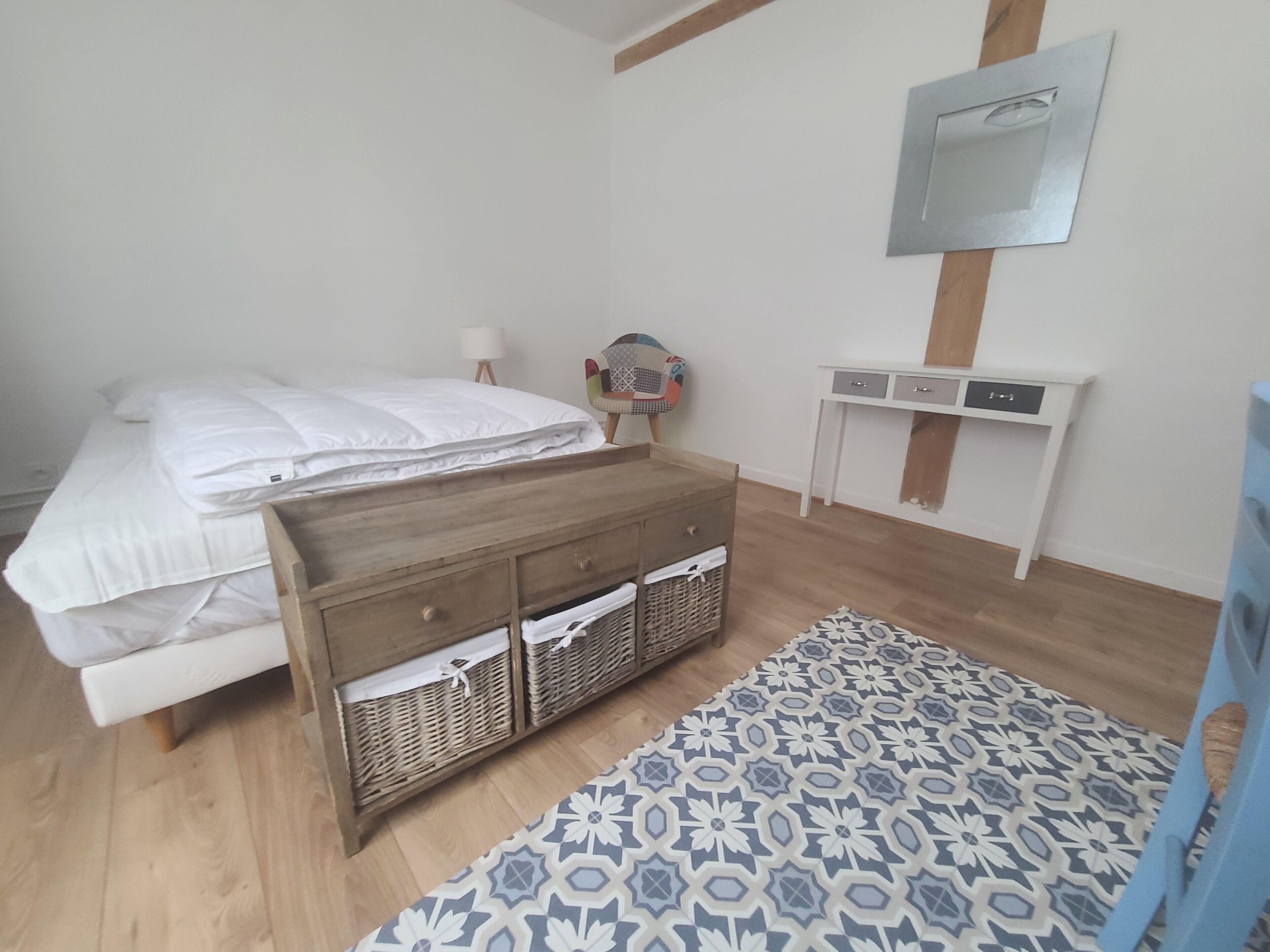 Une chambre à coucher avec un lit, une table de chevet en bois, un petit bureau avec des tiroirs, un panier en osier, un tapis à motifs et un miroir accroché au mur.