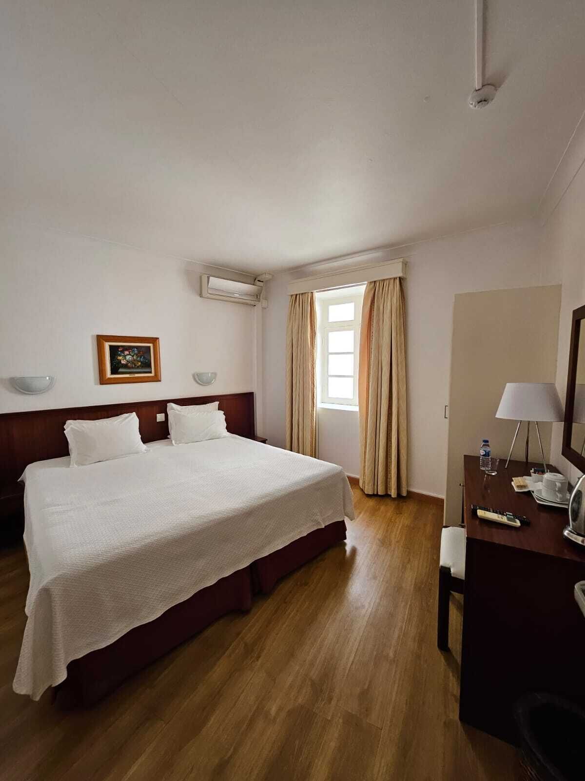 A hotel room with a large bed, a desk with a lamp, a mirror, and a window with curtains.