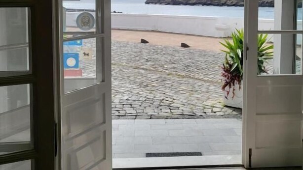 Vista desde un edificio hacia una playa, con un sendero rocoso que lleva a la orilla, una montaña al fondo y una puerta que da a la playa.