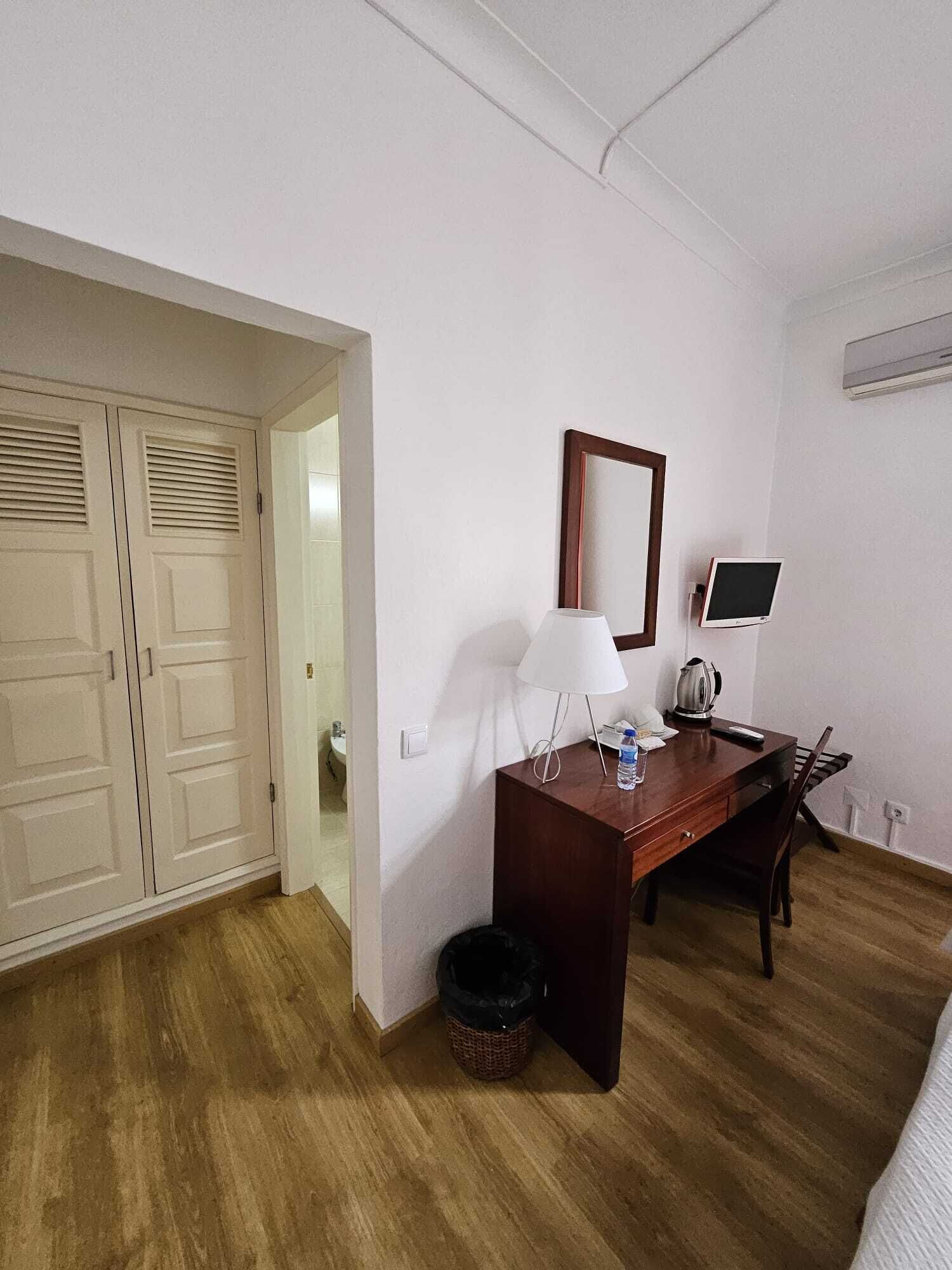 A hotel room with a wooden desk, a chair, a lamp, a small table with a kettle, a trash bin, and a large mirror.