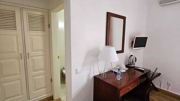 A hotel room with a wooden desk, a chair, a lamp, a small table with a kettle, a trash bin, and a large mirror.