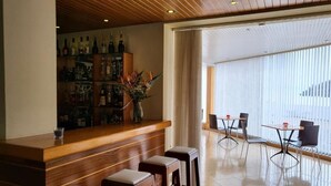 Una zona de bar con mostrador de madera, taburetes y una variedad de botellas. Hay un comedor con una mesa y sillas, y un amplio ventanal con persianas.