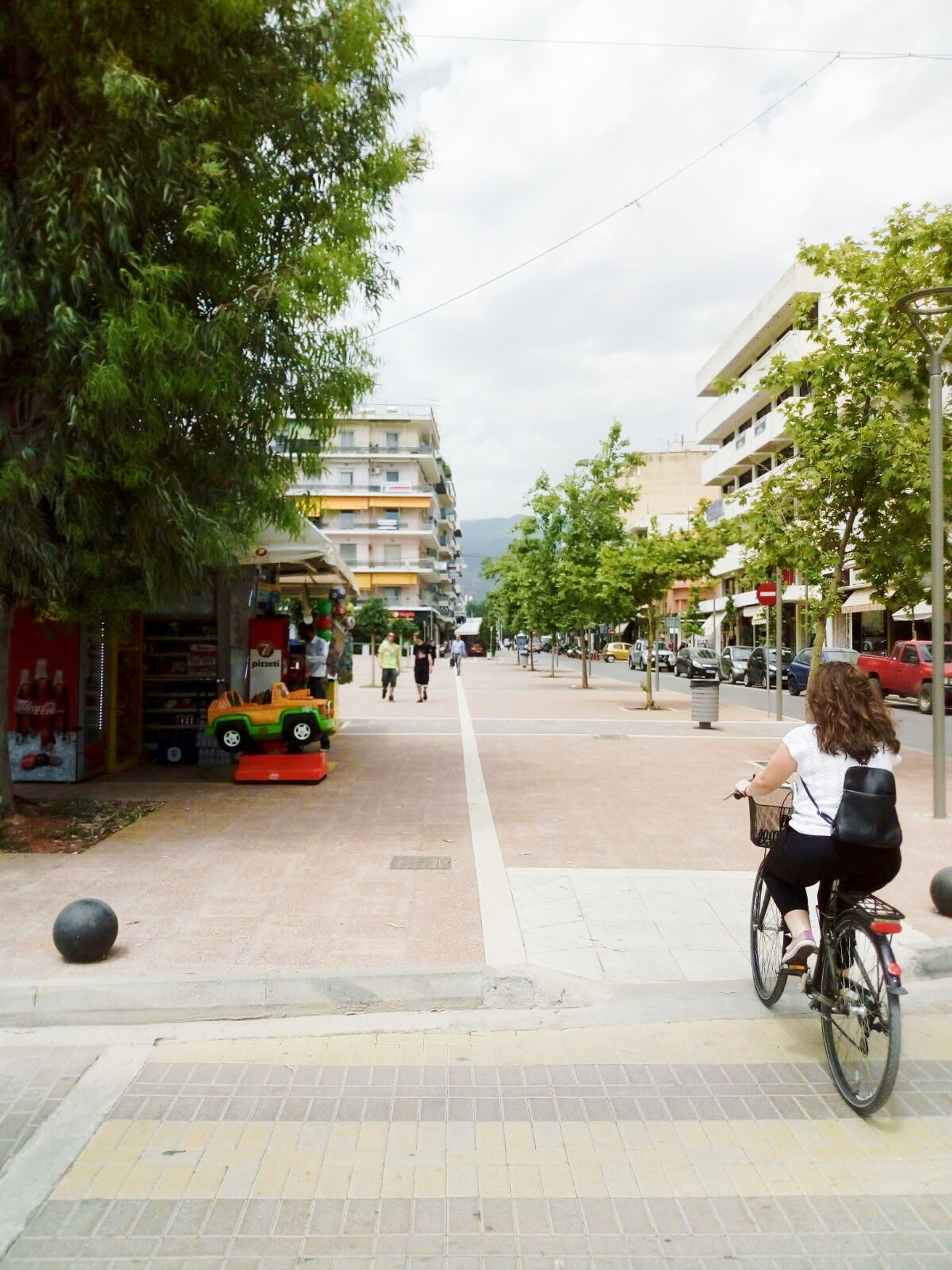 A street scene with a woman riding a bicycle, a toy car, and a building with a sign that reads "7 pizzeria".