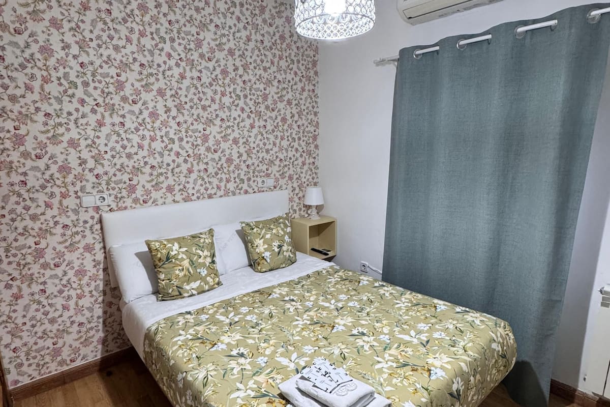 A bedroom with floral wallpaper, a bed with a floral bedspread, a hanging light fixture, an air conditioning unit, and a grey curtain.