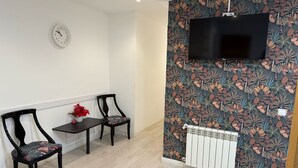A modern living room with a flat-screen TV mounted on a floral wallpapered wall, two dark wooden chairs, a small table with a vase of red flowers, and a white radiator. - Flat55madrid (Madrid)