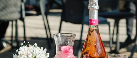 Una mesa con vasos transparentes, una botella y una jarra, probablemente conteniendo bebidas, ubicada al aire libre con sillas y mesas al fondo.
