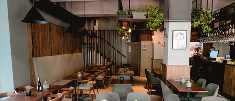 A restaurant interior with wooden tables and chairs, a bar area with wine bottles, and hanging plants.