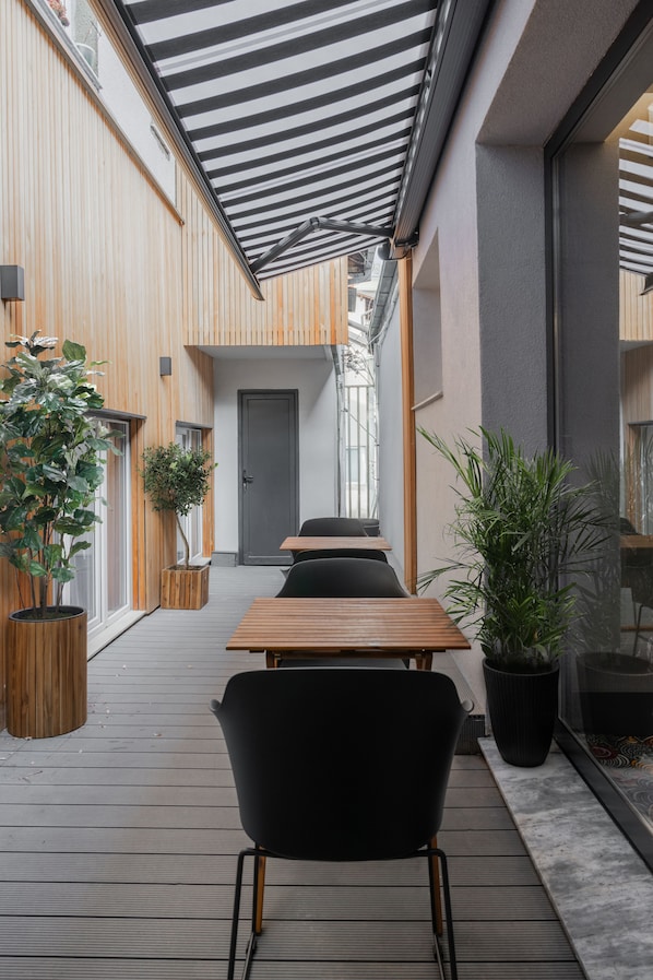 Un área de estar al aire libre moderna con mesas de madera y sillas negras, rodeada de plantas en macetas y un toldo a rayas.