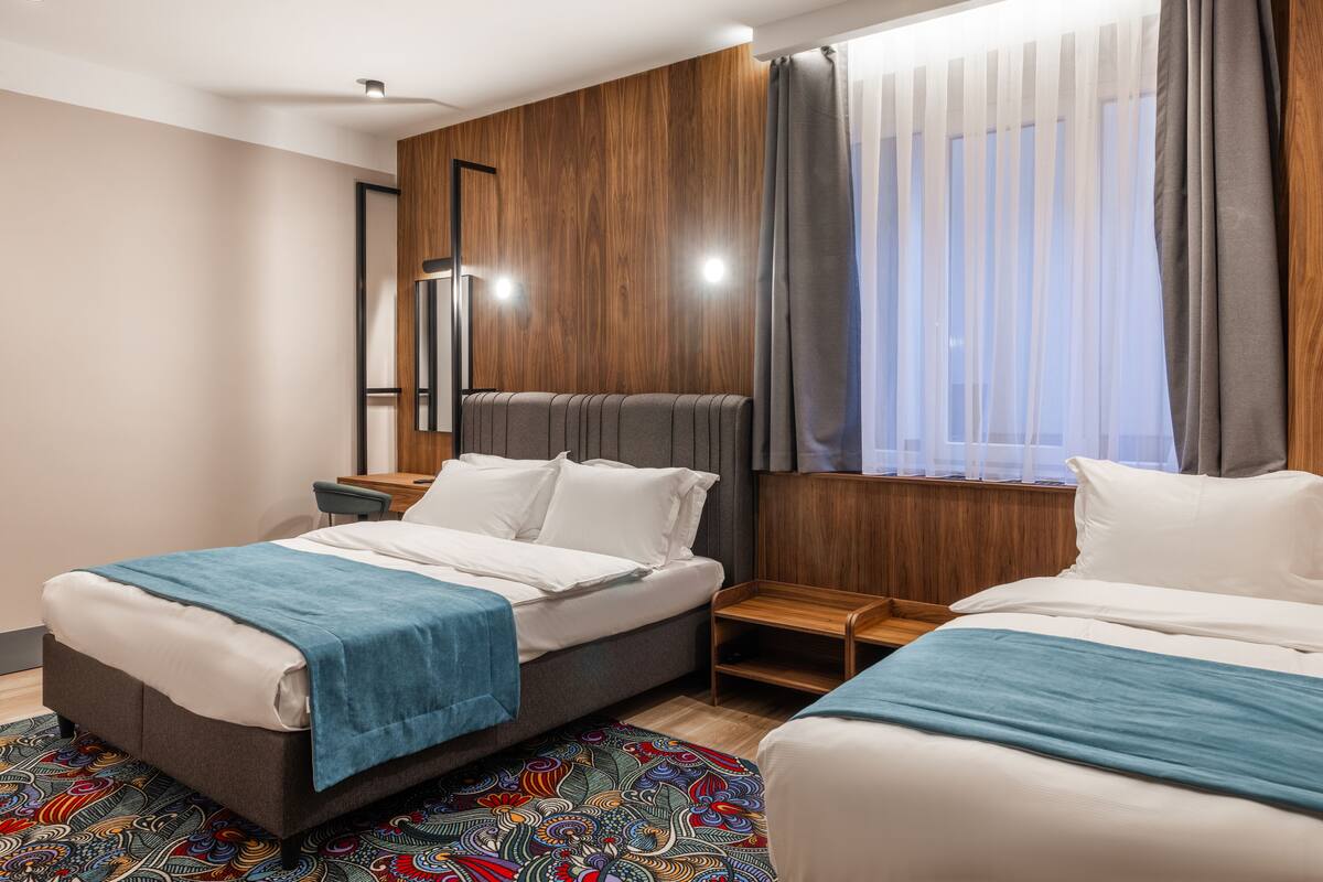 Una habitación de hotel moderna con dos camas, un cabecero de madera, una mesita de noche de madera pequeña y una alfombra colorida con estampado.