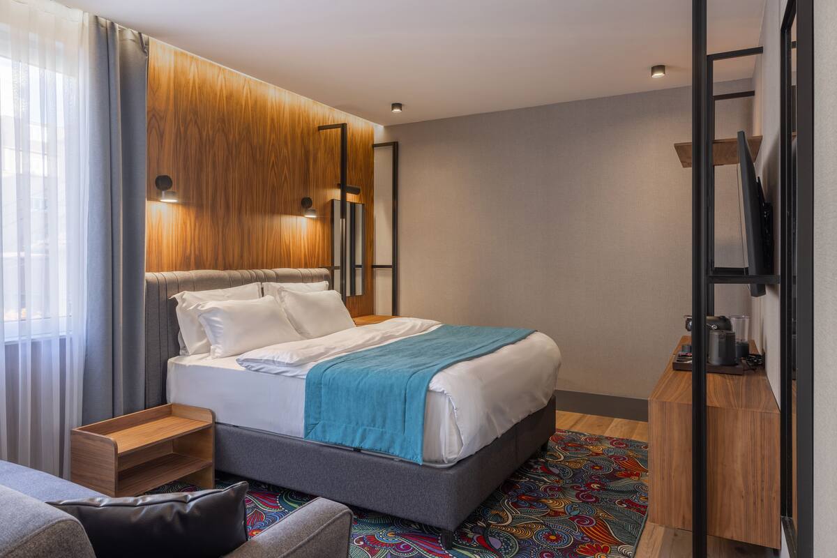Habitación de hotel moderna con una cama grande, un cabecero de madera, una mesita de noche, un pequeño tocador de madera, un tapete colorido con diseño y un mueble empotrado para la televisión.