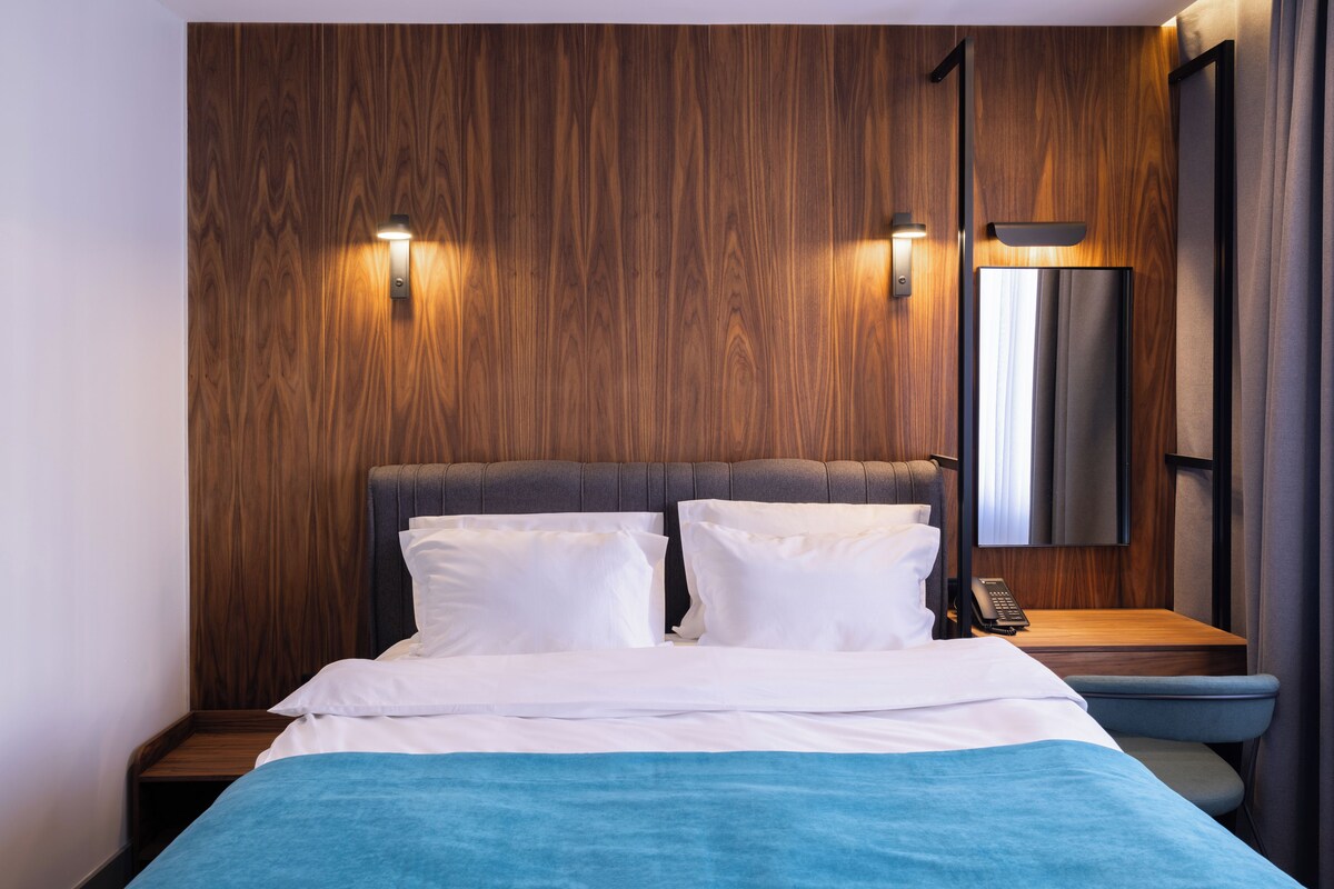 Una habitación de hotel moderna con cabecera de madera, dos mesitas de noche, una cama con ropa blanca y una mesita con una lámpara.