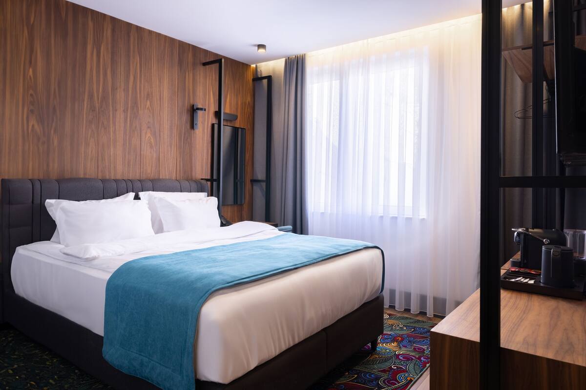 Habitación de hotel con cama, pared de madera, ventana con cortinas y escritorio con cafetera.