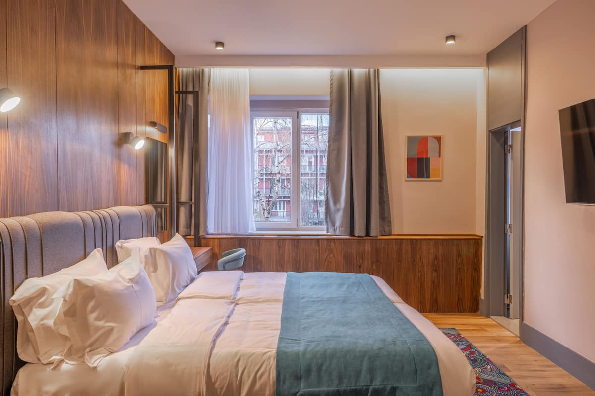 Un dormitorio moderno con una cama grande, paneles de madera, una ventana con cortinas y vistas a edificios.