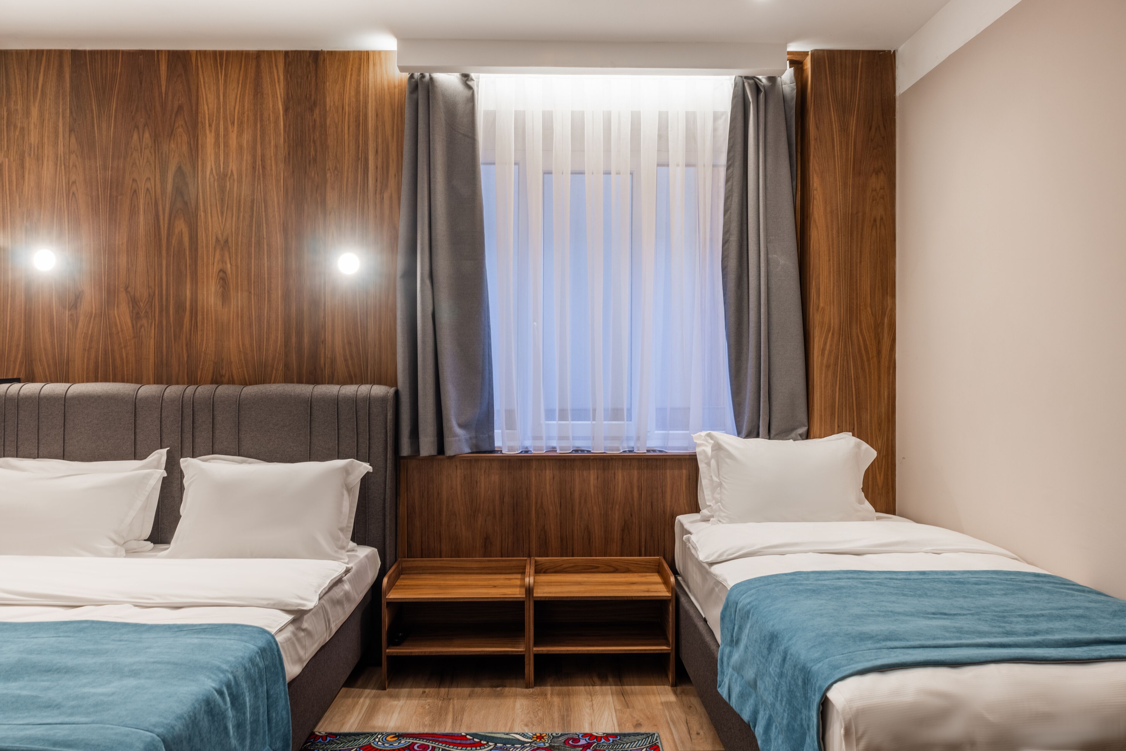 Een hotelkamer met twee bedden, houten hoofdeinden, een raam met dunne gordijnen en een kleine houten tafel tussen de bedden.