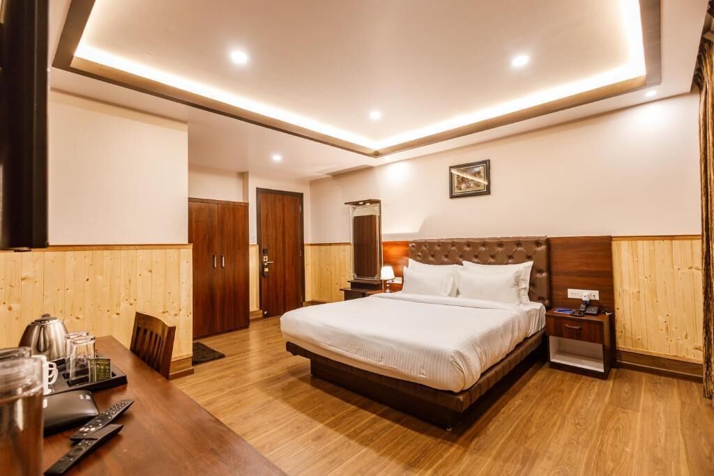 Habitación de hotel con una cama grande, suelo de madera, una mesa de comedor con sillas, una pequeña mesa con hervidor y una mesita de noche con lámpara.