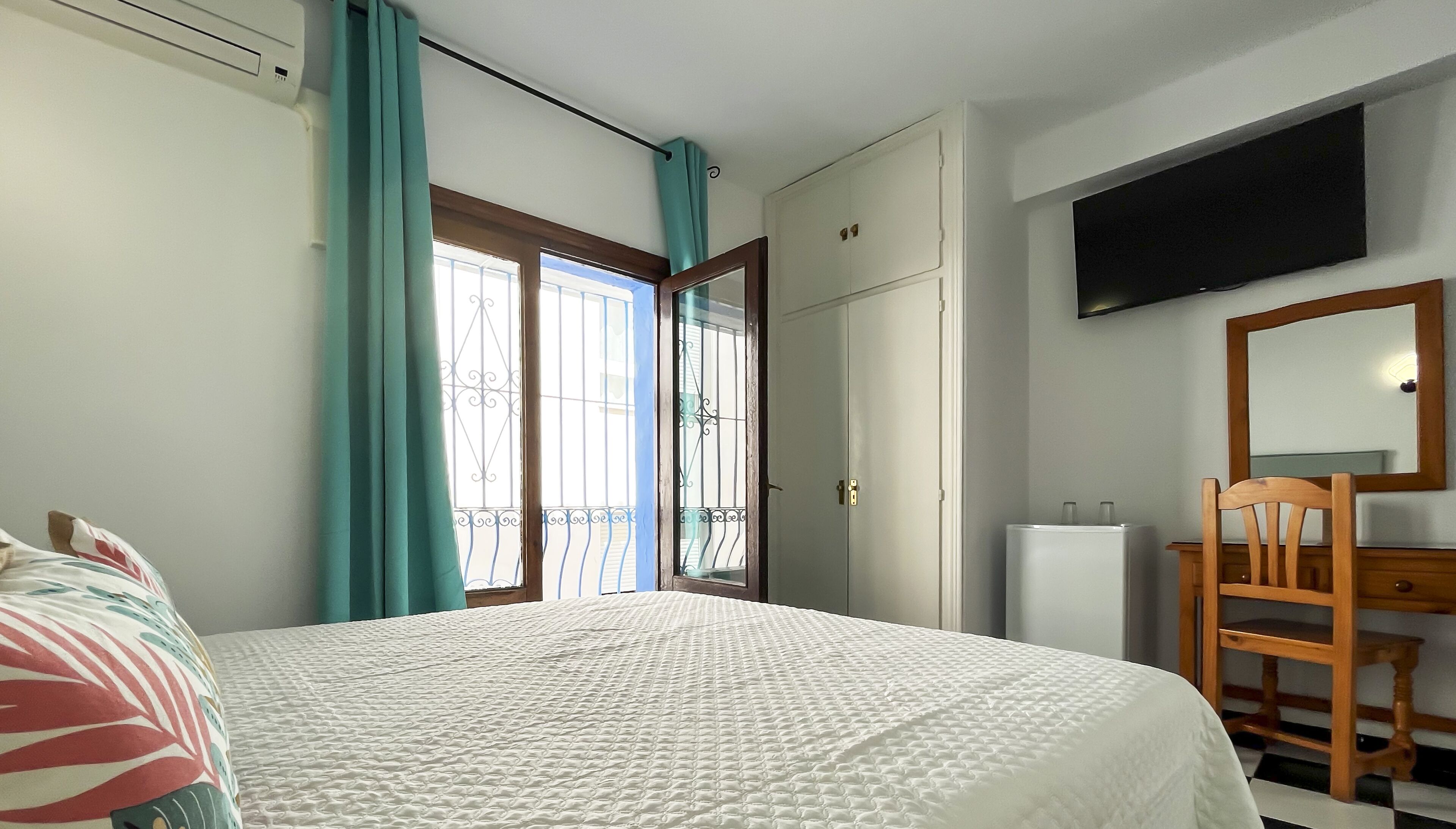 Une chambre à coucher avec un lit, une chaise en bois, une commode, un téléviseur et une fenêtre avec des rideaux.