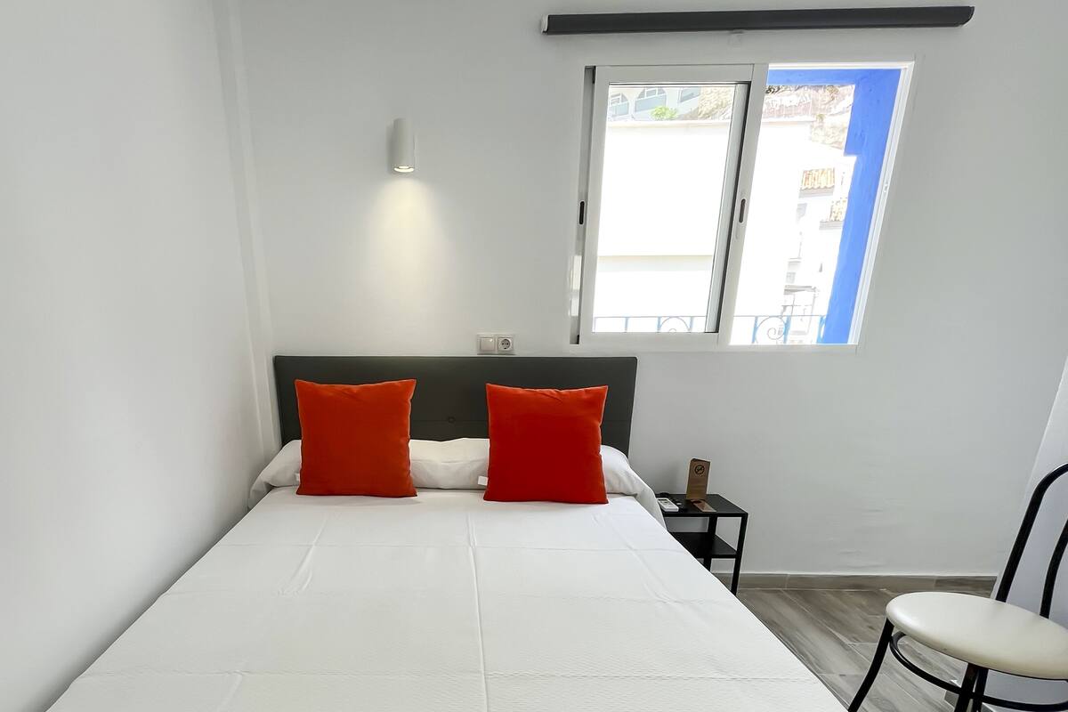 Ein Schlafzimmer mit einem Bett, zwei roten Kissen, einem Nachttisch, einem Stuhl und einem Fenster mit Blick auf Gebäude.