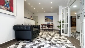 A modern interior with a black leather sofa, a dining area with wooden chairs, a tiled floor, and a large window with a view of the sea. - Micaela Charming Hostal (Torremolinos)
