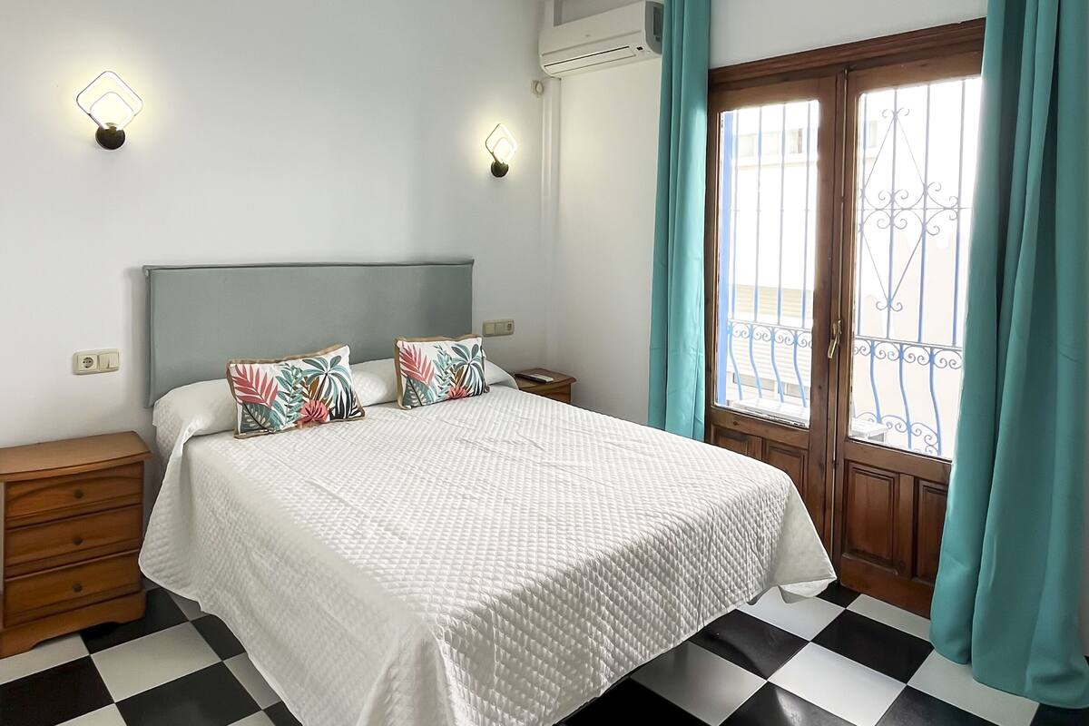 Ein Schlafzimmer mit Schachbrettmuster-Boden, einem Bett mit weißer Steppdecke, zwei Nachttischen, einem Holz-Kopfteil und einem Fenster mit türkisfarbenen Vorhängen.