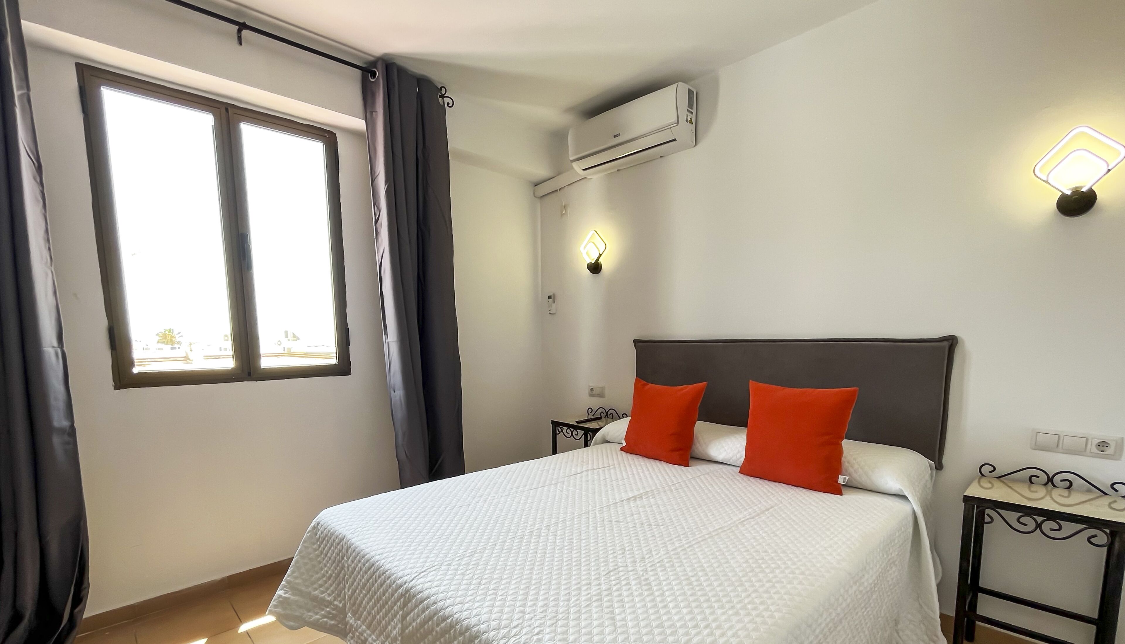 A hotel room with a bed, a nightstand, an air conditioning unit, a wall-mounted light, and a window with curtains.