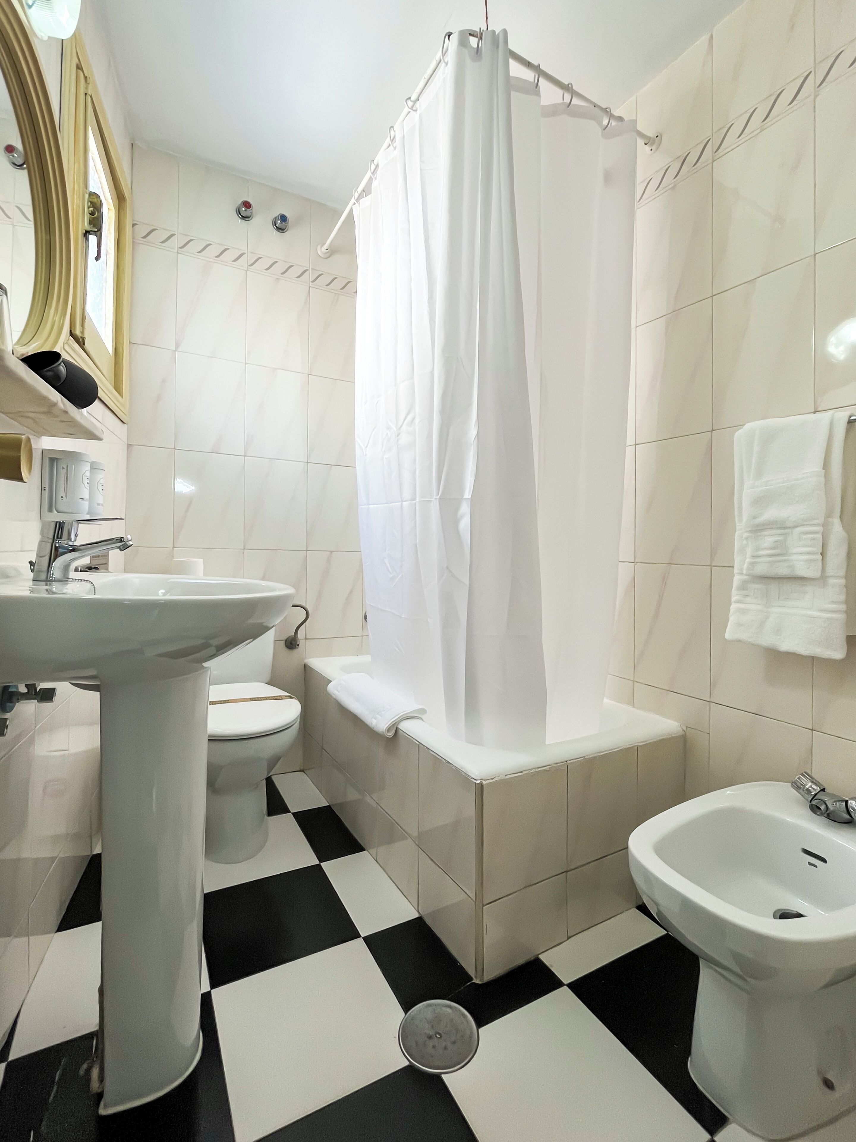 Un baño con lavamanos, inodoro y bidé blancos, cortina de ducha y piso a cuadros.