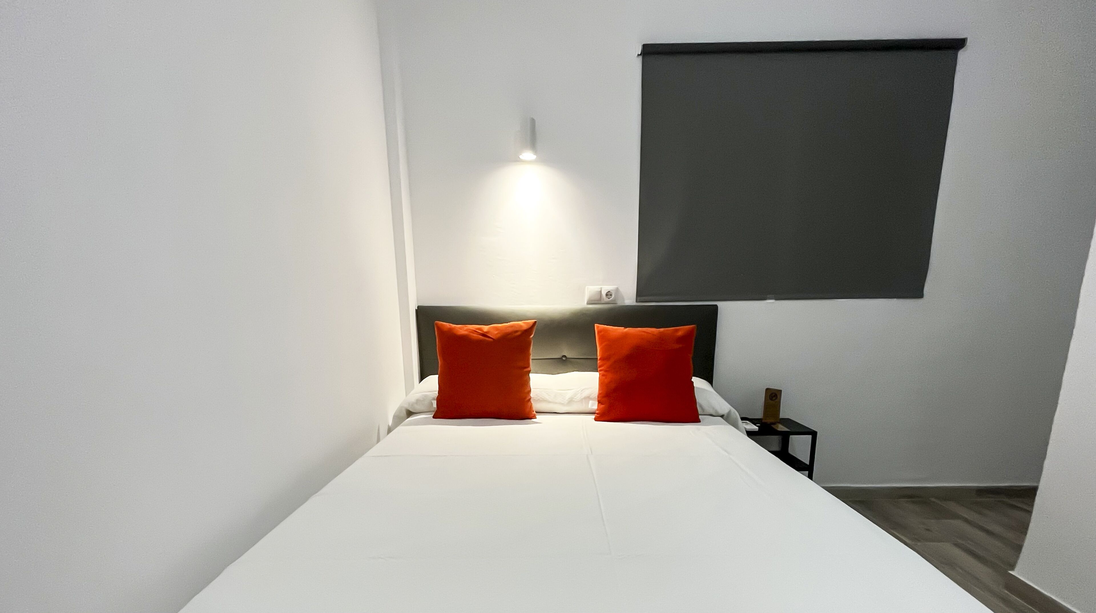 A small, neatly made bed with white bedding and two orange pillows, a bedside table with a lamp, and a dark-colored window blind.