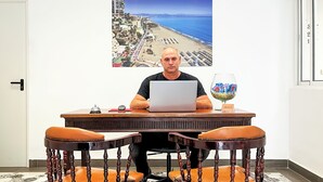 A man sitting at a wooden table with a laptop, two wooden chairs with orange cushions, a glass of water, and a framed picture of a beach and buildings on the wall. - Micaela Charming Hostal (Torremolinos)