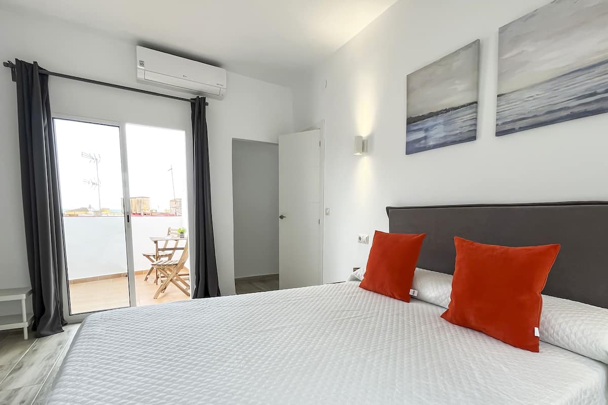 Ein Hotelzimmer mit einem großen Bett, zwei roten Kissen, einem grauen Kopfteil und Blick ins Freie durch eine Schiebetür.