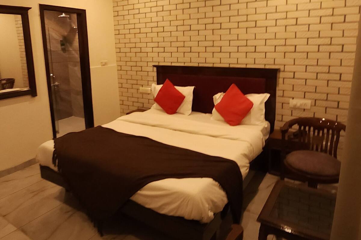 A bedroom with a brick-patterned wall, a bed with white bedding and red pillows, a ceiling fan, an air conditioning unit, a wooden chair, and a small table.