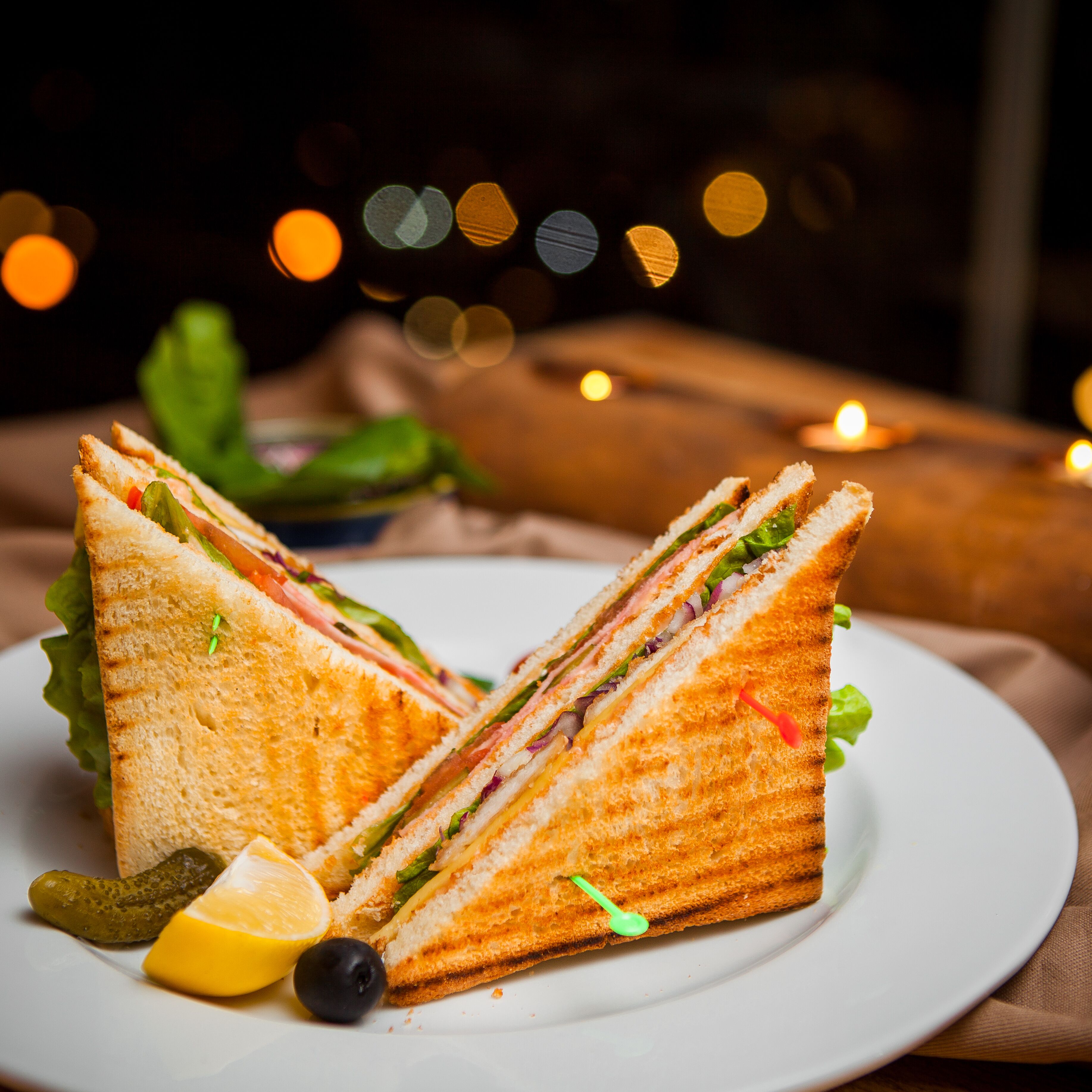 Un sandwich servi sur une assiette, accompagné d’une portion de cornichons, d’un quartier de citron et d’une olive.