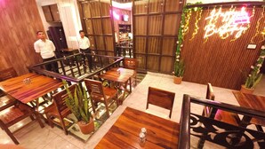 Un restaurante con mesas y sillas de madera, barandilla de vidrio y plantas en macetas.