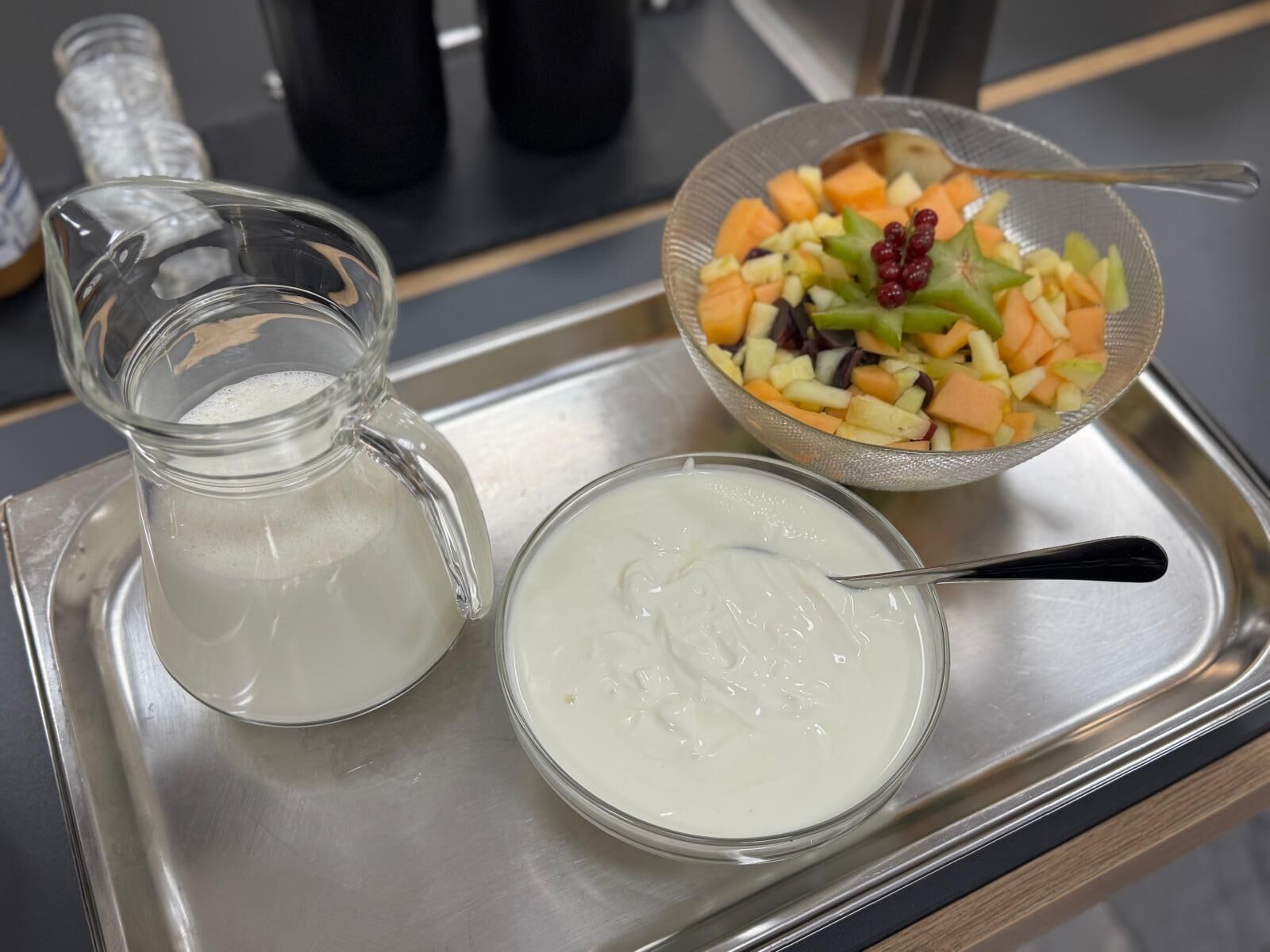 Una bandeja con una jarra de líquido transparente, un cuenco de ensalada de frutas variadas y un recipiente con sustancia blanca cremosa.
