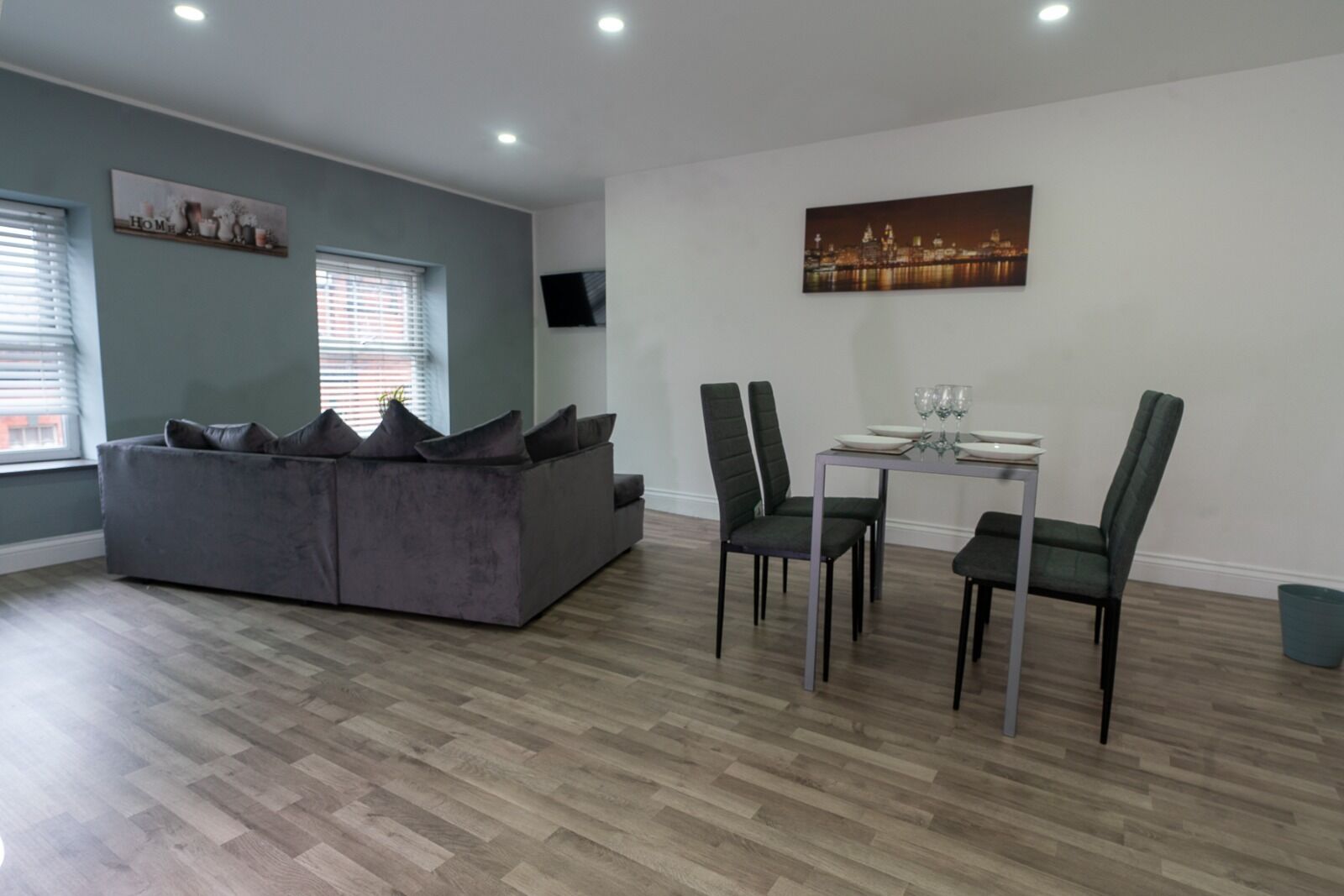 A modern living room with a grey sofa, dining table with chairs, and a cityscape painting on the wall.