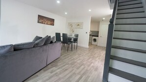 Un salon moderne avec un canapé gris, un coin repas avec des chaises, une cuisine équipée d’un lave-linge et un escalier.