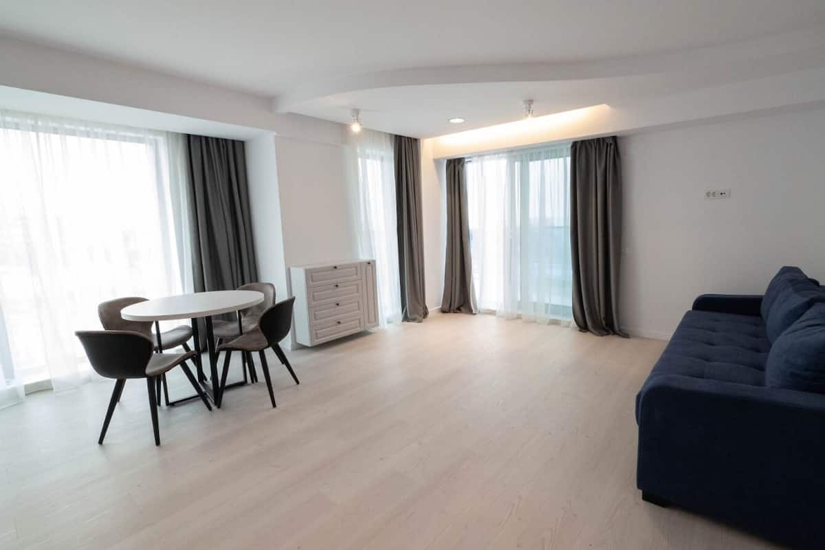 A spacious living area with a dark blue sofa, a round dining table with chairs, and large windows with curtains.