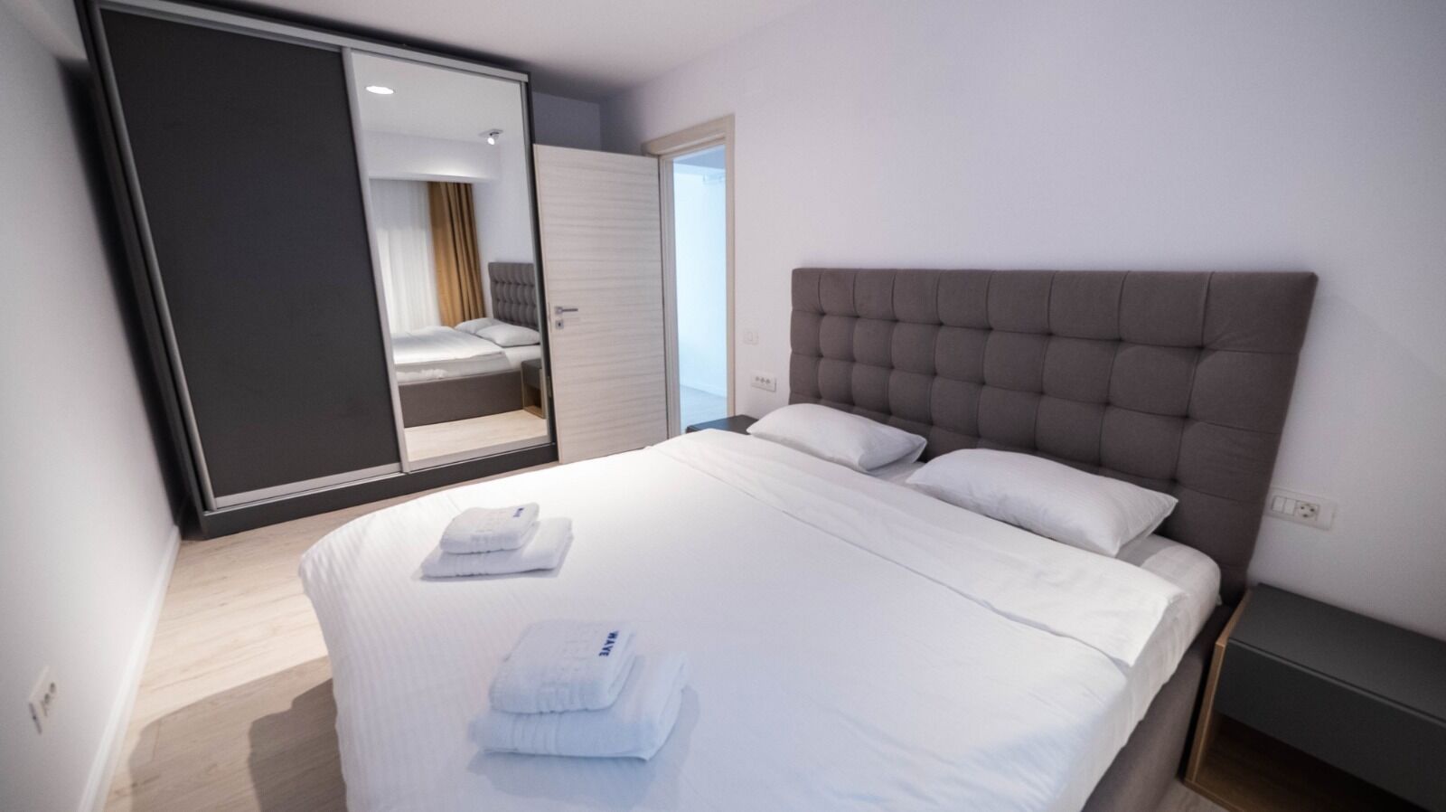 Une chambre d’hôtel moderne avec un grand lit, une tête de lit rembourrée de couleur grise et une porte coulissante donnant sur un balcon.
