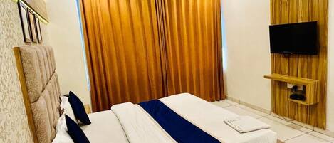 A hotel room with a bed, a wooden headboard, a television mounted on a wooden panel, and a window with curtains.
