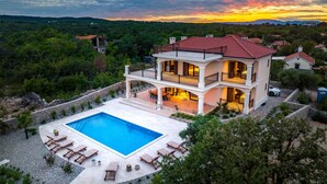 Ein zweistöckiges Haus mit Swimmingpool, umgeben von Bäumen und einem Sonnenuntergang im Hintergrund.