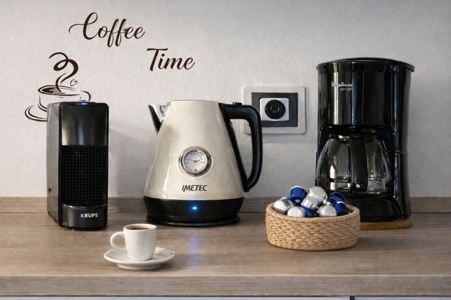 Een koffiehoek met een koffiezetapparaat van Krups, een waterkoker van IMETEC, een koffiekan en een mand met koffiecups.