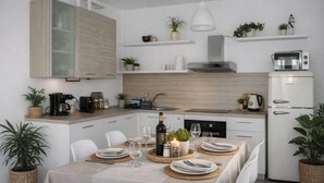 Ett modernt kök med vita skåp, en inbyggd spis och ett vitt kylskåp. Det finns växter på bänkskivorna och hyllorna, och ett matbord för två personer med tallrikar, glas och en flaska vin.