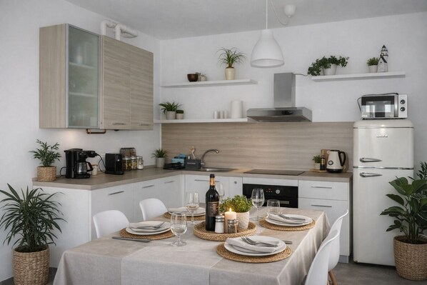 A modern kitchen with white cabinetry, a built-in stove, and a white refrigerator. There are plants on the countertops and shelves, and a dining table set for two with plates, glasses, and a bottle of wine. - Lago Maggiore holiday house (Dumenza)